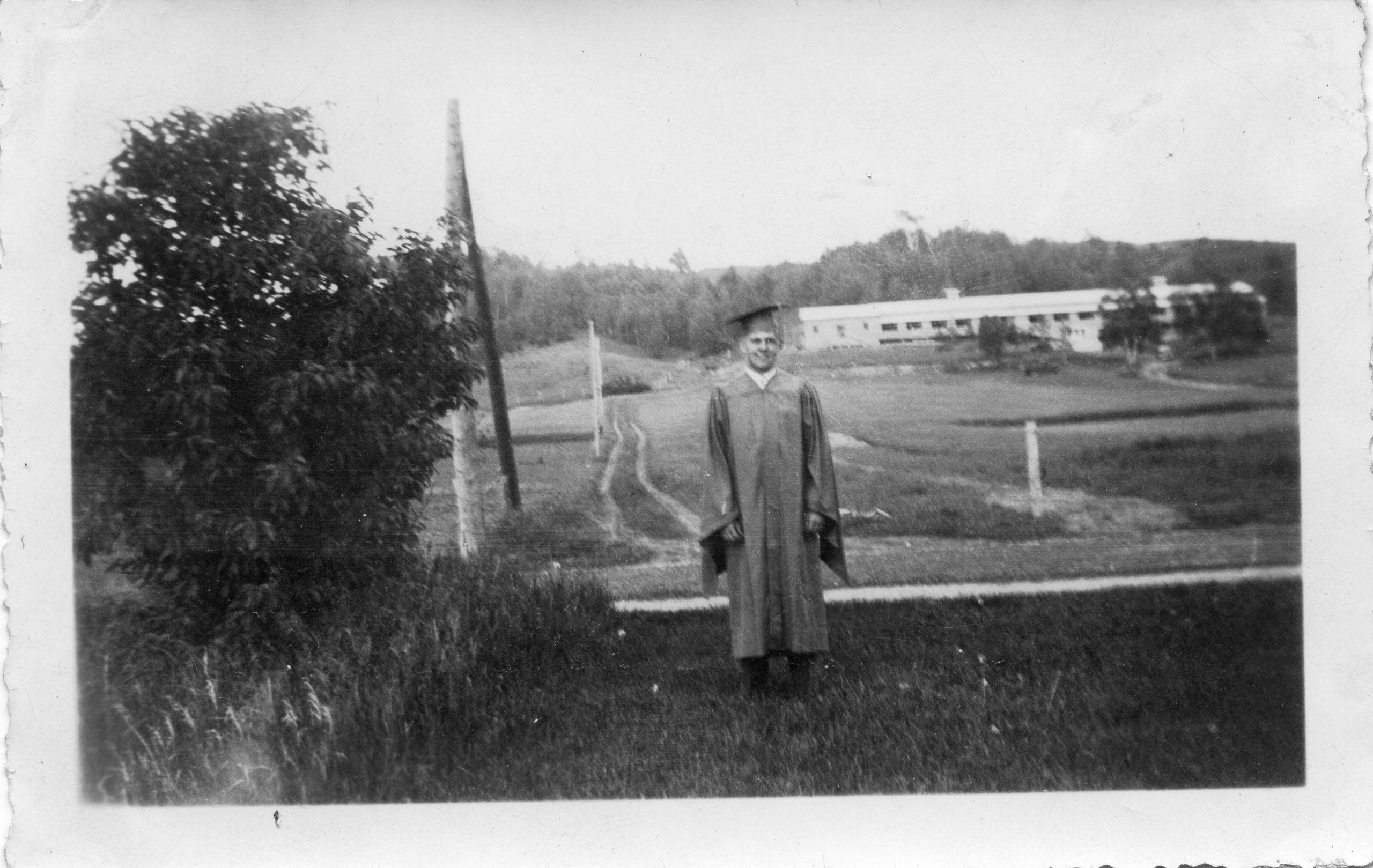 Robert Peacock in a graduation cap and gown, in front of a rural scene.